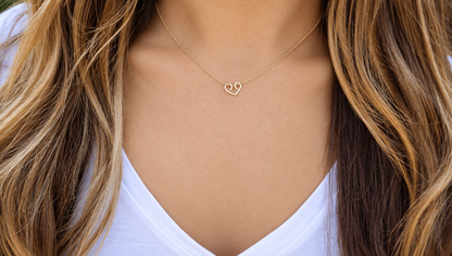 A close-up shot of a model wearing a dainty, handmade gold looped wire heart necklace on a delicate chain against her collarbone.
