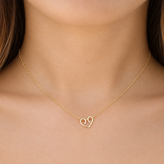 A close-up shot of a model wearing a dainty, handmade gold looped wire heart necklace on a delicate chain against her collarbone.