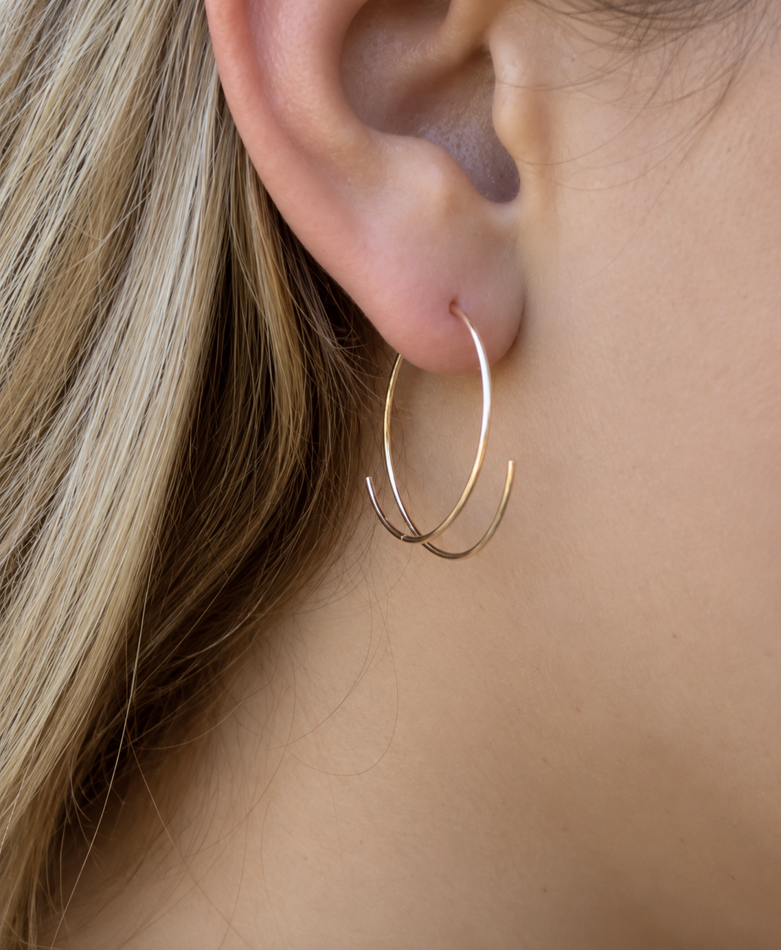 A close-up of a person's ear wearing a delicate, thin gold hoop earring with a second, larger gold hoop layered behind it.