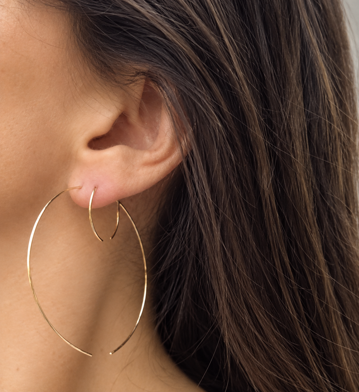 A close-up of a person's ear wearing a pair of large gold hoop earrings, with dark brown hair visible against a neutral light gray background.