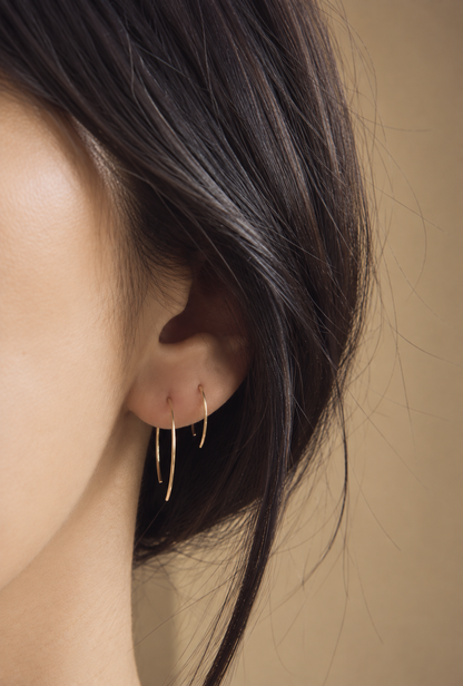 A close-up of a model's ear wearing a pair of delicate gold hoop earrings against a warm beige background.