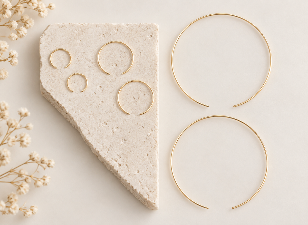 Four gold-toned open hoop earrings displayed on a light neutral surface, with three hoops arranged on a textured stone slab and one larger hoop on the right. Delicate dried flowers appear along the left edge.