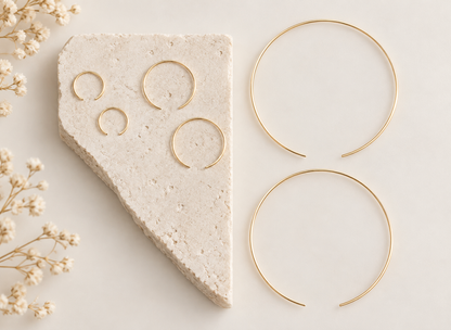 Four gold-toned open hoop earrings displayed on a light neutral surface, with three hoops arranged on a textured stone slab and one larger hoop on the right. Delicate dried flowers appear along the left edge.