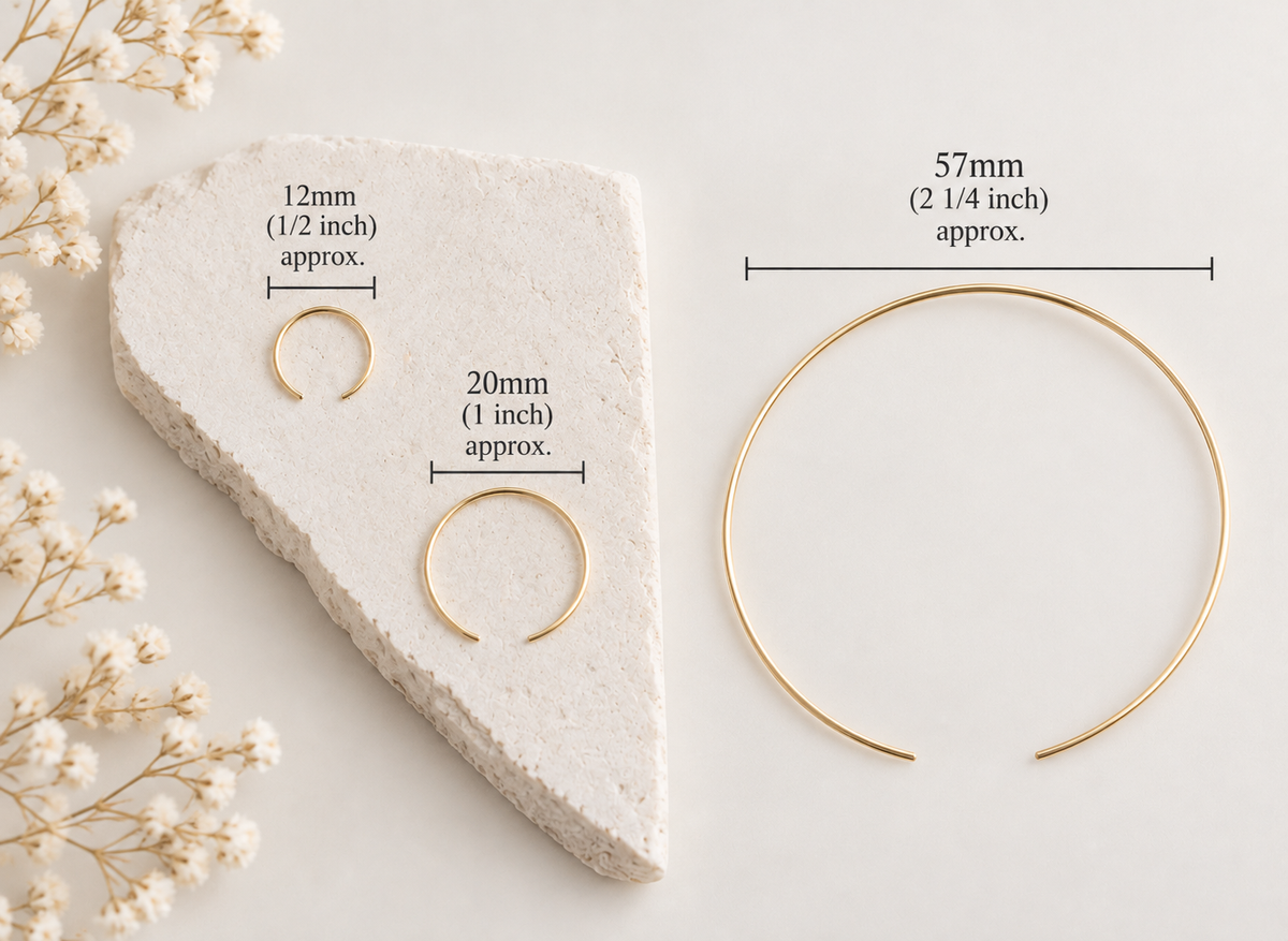 Three gold-toned open hoop or ring-style jewelry pieces displayed on a light neutral background with overlaid size labels: 12mm (1/2 inch), 20mm (1 inch), and 57mm (2 1/4 inch). A textured stone slab and small dried flowers are styling props.
