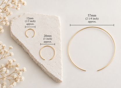 Three gold-toned open hoop or ring-style jewelry pieces displayed on a light neutral background with overlaid size labels: 12mm (1/2 inch), 20mm (1 inch), and 57mm (2 1/4 inch). A textured stone slab and small dried flowers are styling props.