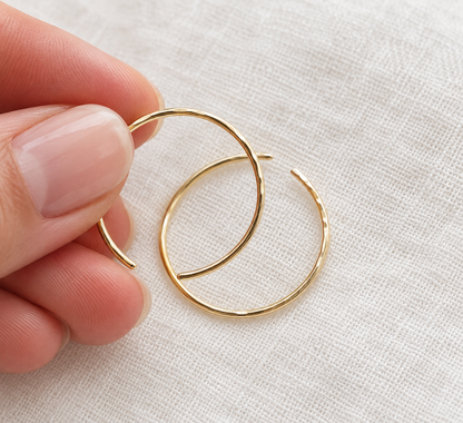 A close-up of a hand holding a pair of thin, hammered gold hoop earrings against a textured off-white fabric background.